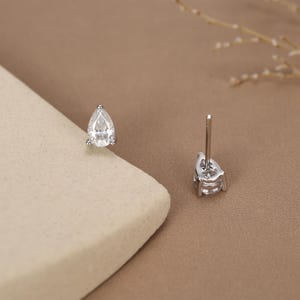 May include: A pair of silver stud earrings with pear-shaped, clear gemstones. The earrings are set in a silver metal and have a post backing. The background is a neutral tan color.