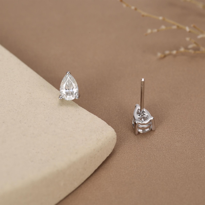 May include: A pair of silver stud earrings with pear-shaped, clear gemstones. The earrings are set in a silver metal and have a post backing. The background is a neutral tan color.