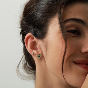 Puede incluir: Un par de pendientes de bot&oacute;n con una peque&ntilde;a polilla verde de alas delicadas y una diminuta piedra transparente facetada situada encima. El pendiente de polilla tiene un acabado de esmalte verde vibrante con bordes plateados.