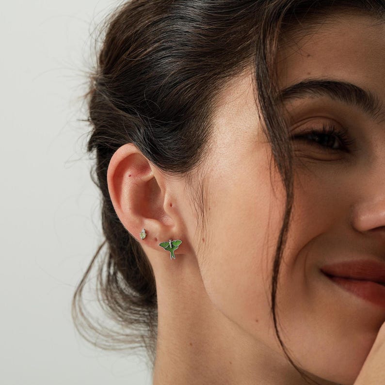 Puede incluir: Un par de pendientes de bot&oacute;n con una peque&ntilde;a polilla verde de alas delicadas y una diminuta piedra transparente facetada situada encima. El pendiente de polilla tiene un acabado de esmalte verde vibrante con bordes plateados.