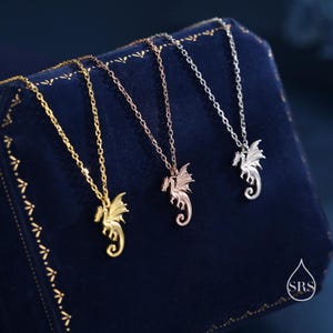 May include: Three delicate necklaces featuring small dragon pendants in gold, rose gold, and silver tones. Each pendant is suspended from a matching fine link chain. A small white logo with the letters SRS inside a droplet shape is visible.