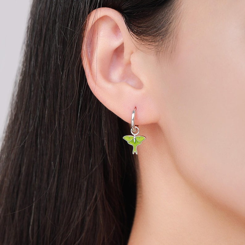 May include: A silver hoop earring with a lime green moth charm. The moth charm has black detailing and is enameled. The earring is shown on a person's ear.