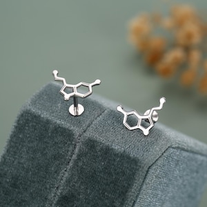 Op de afbeelding: Een paar zilverkleurige oorstekers in de vorm van een serotonine-molecuul. Eén oorbel is een oorsteker met pin, de andere heeft een platte achterkant. Ze worden gepresenteerd op een donkergroen fluwelen oppervlak. Wetenschappelijk accessoire.