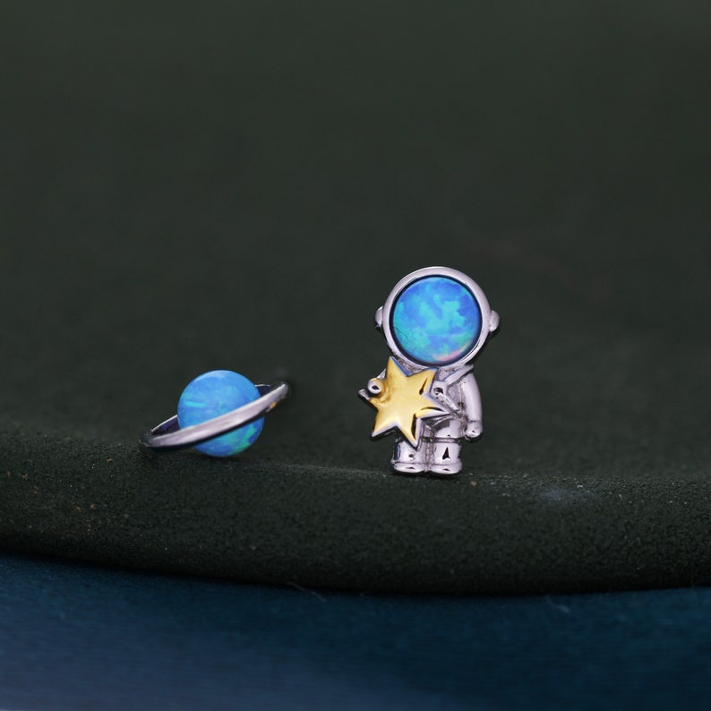 May include: Silver-toned astronaut and planet earrings. The astronaut holds a gold star, and the planet has a silver ring. Both have blue opal-like centers. The earrings are set against a dark green background.