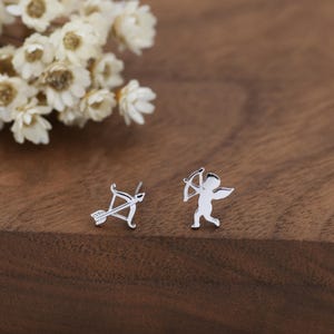 May include: A pair of silver stud earrings. One earring features a bow and arrow, and the other depicts a small cupid figure with a bow. The earrings are on a wooden surface with white flowers in the background.