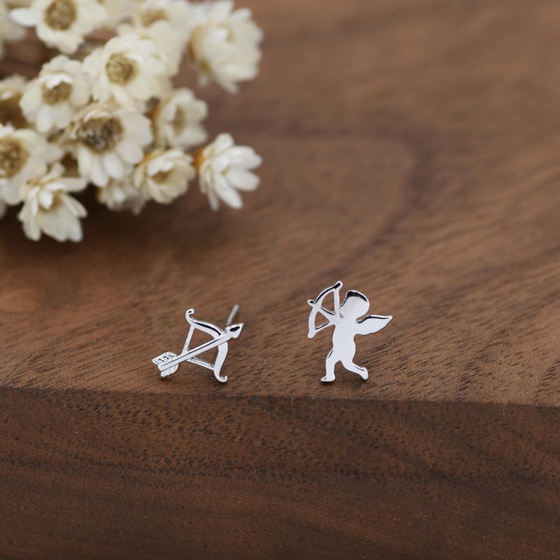 May include: A pair of silver stud earrings. One earring features a bow and arrow, and the other depicts a small cupid figure with a bow. The earrings are on a wooden surface with white flowers in the background.