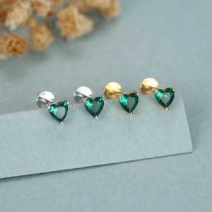 May include: Four stud earrings with heart-shaped, emerald-green gemstones. Two earrings have silver posts, and two have gold posts. The earrings are displayed on a light blue surface, with a blurred background of dried flowers.