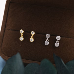 Pode incluir: Três pares de brincos pendurados com pedras de zircônia cúbica transparentes. Os brincos estão sobre um fundo de veludo marrom.