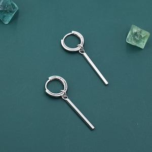 May include: A pair of silver hoop earrings with a minimalist design. Each earring features a small hoop with a straight, rectangular bar pendant. The earrings are set against a dark teal background.