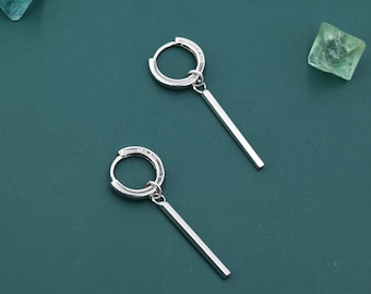 Puede incluir: Un par de pendientes de aro plateados con un diseño minimalista. Cada pendiente presenta un pequeño aro con una barra rectangular recta. Los pendientes se muestran sobre un fondo verde oscuro.