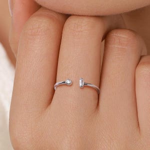 Puede incluir: Un anillo abierto de plata con dos piedras preciosas transparentes. Una piedra es redonda, la otra rectangular. El anillo está en un dedo.