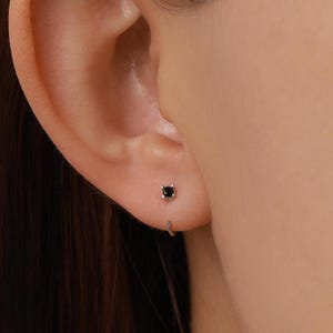Può includere: Un piccolo orecchino color argento con una singola pietra preziosa nera rotonda incastonata in una montatura a quattro griffe. L'orecchino è progettato come un delicato cerchio che attraversa il lobo, con una finitura metallica lucida.
