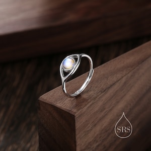 May include: A silver ring featuring a round, iridescent gemstone within an open, eye-like frame. The adjustable ring is displayed on a dark wooden surface. The gemstone exhibits a spectrum of colours, including blue, yellow, and white.