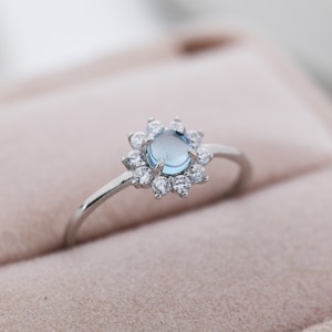 Puede incluir: Un anillo de plata con una piedra preciosa azul claro rodeada de piedras preciosas más pequeñas y transparentes en forma de flor.