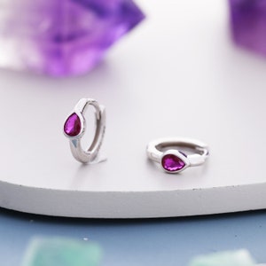 May include: A pair of silver hoop earrings with a small teardrop-shaped pink gemstone set in each hoop.
