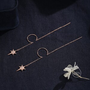 May include: A pair of rose gold earrings with a star charm and a hoop. The earrings are on a dark blue fabric background.