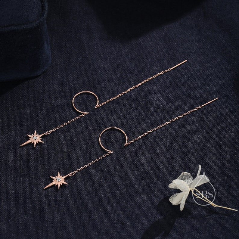 May include: A pair of rose gold earrings with a star charm and a hoop. The earrings are on a dark blue fabric background.