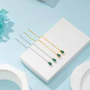 May include: Four pairs of threader earrings with teardrop-shaped green gemstones. Two pairs are silver-toned, and two are gold-toned. The earrings are displayed on a white surface against a light blue background, with a touch of floral decor.