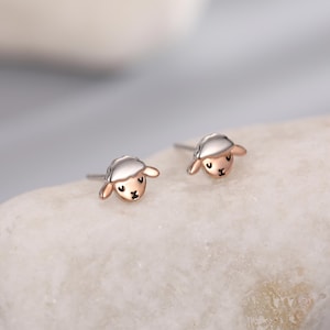 May include: A pair of stud earrings with a sheep design. The earrings have a silver-coloured head and a rose gold-coloured face and ears. The sheep's face has a small, black nose and eyes. The earrings are small and delicate.
