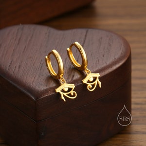 May include: A pair of gold hoop earrings with a dangling eye of Horus charm. The earrings are on a wooden surface.