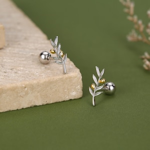 Peut inclure: Une paire de boucles d'oreilles à tige argentées arborant un motif de branche d'olivier avec de petits accents dorés polis représentant des olives. Chaque boucle possède un fermoir à bille et un motif de feuilles texturé et délicat.