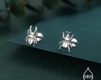 May include: A pair of silver-colored stud earrings, each shaped like a beetle. The earrings are detailed with legs, antennae, and a textured body. The earrings are displayed on a dark green surface.