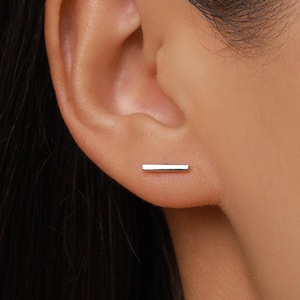 May include: A small, polished silver-tone bar stud earring positioned horizontally on the earlobe. The earring features a clean, rectangular shape with a smooth, reflective metallic surface that catches the light, creating a simple and understated appearance.