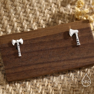 May include: A pair of silver-colored axe-shaped stud earrings. The earrings are displayed on a dark wooden surface, with a neutral-toned woven background. The axes have detailed handles and blades, showcasing a miniature design.