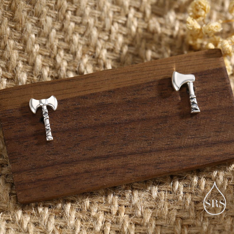 May include: A pair of silver-colored axe-shaped stud earrings. The earrings are displayed on a dark wooden surface, with a neutral-toned woven background. The axes have detailed handles and blades, showcasing a miniature design.