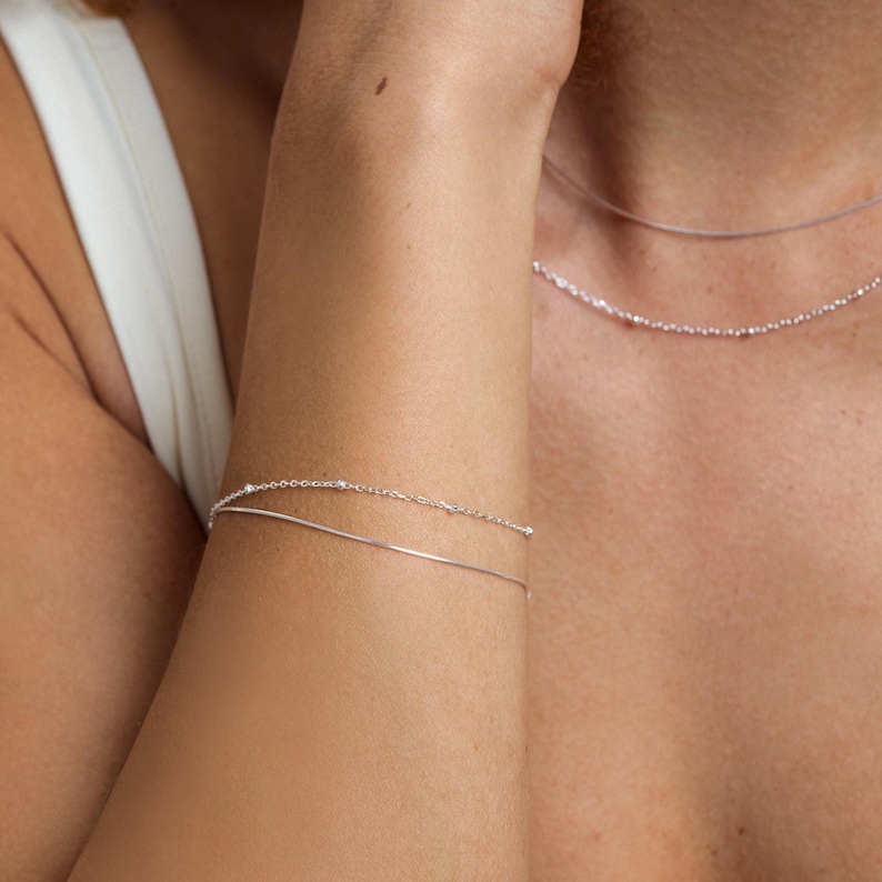 May include: A double-layered silver bracelet consisting of one delicate plain chain and one satellite chain with small evenly spaced beads. The bracelet is worn on the wrist, complementing a matching silver necklace visible around the neck area.
