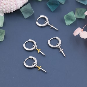 Könnte beinhalten: Drei Paar silberne Creolen mit einem kleinen Schwertanhänger, der an jedem Creolen hängt. Die Schwerter sind goldfarben.