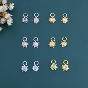 Pode incluir: Uma coleção de pingentes em forma de sol em tons de prata e ouro, cada um com uma pedra central iridescente ou opalescente. Os pingentes estão dispostos sobre um fundo verde-azulado escuro, mostrando seus detalhes intrincados e acabamentos variados.