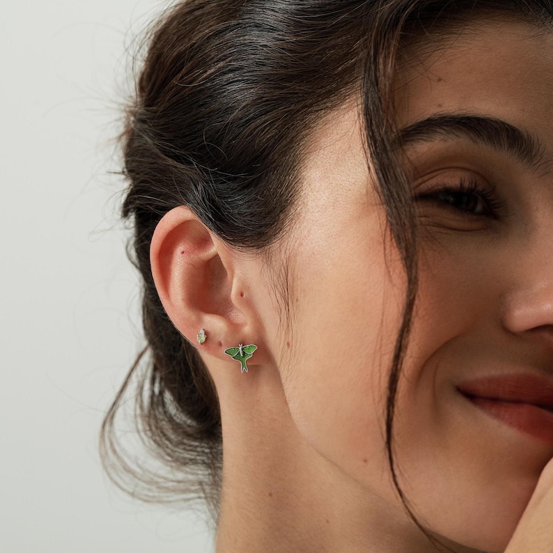 May include: A pair of small stud earrings consisting of one clear gemstone stud and one green moth-shaped stud. The moth earring features detailed wings with a light green center and darker green edges, while the other is a simple faceted stone.