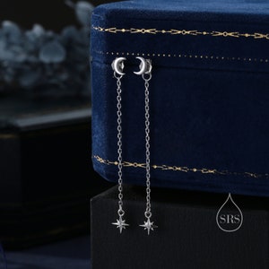 Puede incluir: Un par de pendientes de plata con un dije de luna creciente y un dije de estrella colgante. Los pendientes se muestran en una caja de terciopelo azul.