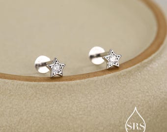 Puede incluir: Dos pendientes plateados en forma de estrella con piedras preciosas transparentes. Los pendientes están sobre una superficie marrón claro. Cada pendiente tiene un pequeño respaldo redondo. Las estrellas tienen un diseño de cinco puntas con una piedra preciosa en el centro.
