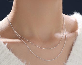 May include: A layered silver necklace featuring two distinct chains. The top chain is a smooth, thin snake chain, while the bottom chain is a delicate, fine-linked cable chain. Both chains are metallic silver in colour and rest against the neck.