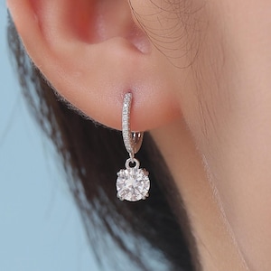 May include: A silver hoop earring with a dangling, round, clear gemstone. The hoop is encrusted with small, clear stones. The earring is shown on a person's ear.