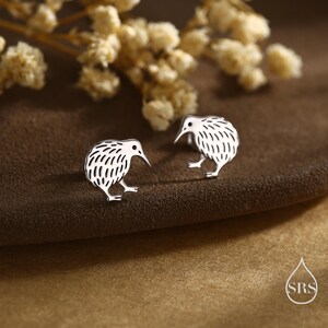 May include: A pair of silver-colored kiwi bird stud earrings. Each earring features a detailed kiwi bird design with a textured body and a long beak. The earrings are displayed on a brown surface with dried flowers in the background.