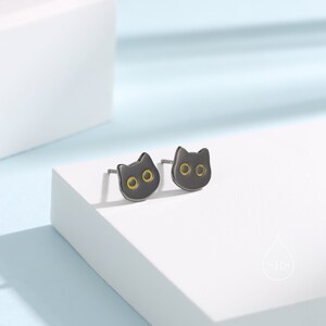Peut inclure: Une paire de boucles d'oreilles clous en forme de chat noir avec des yeux jaunes. Les boucles d'oreilles sont sur une surface blanche.