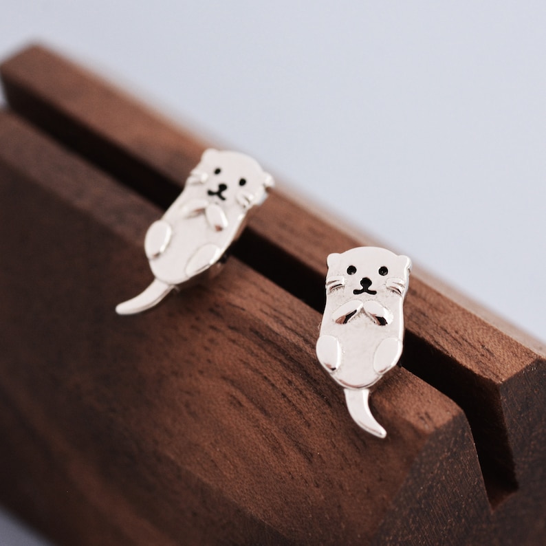 May include: A pair of silver stud earrings featuring a cute otter design. The otters are facing forward with their paws clasped in front of them.