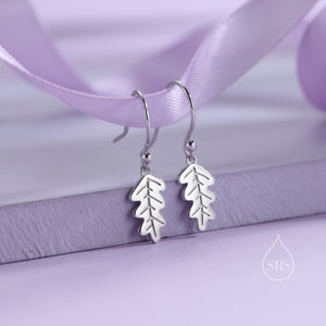 Puede incluir: Pendientes de plata con diseño de hoja. Los pendientes están sobre una superficie morada con una cinta morada.