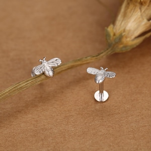 May include: A pair of silver-toned bee stud earrings featuring detailed wings and a striped body. One earring is positioned on a thin stem, while the other stands upright on its flat circular backing, showcasing the post and secure closure mechanism.