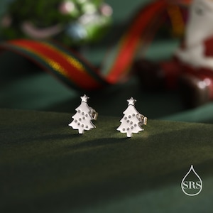 Könnte beinhalten: Ein Paar silberne Weihnachtsbaum-Ohrstecker. Jeder Ohrring hat die Form eines Baumes mit einem Stern und Punktdetails. Die Ohrringe sind vor einem grünen Hintergrund mit verschwommenen Weihnachtsdekorationen platziert.