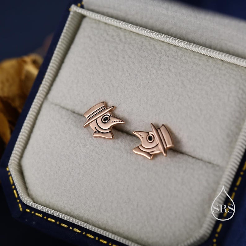 May include: A pair of rose gold-colored stud earrings shaped like plague doctor masks. The earrings are displayed inside a blue jewelry box with a light gray interior. The earrings have a hat, a long beak, and eye details.