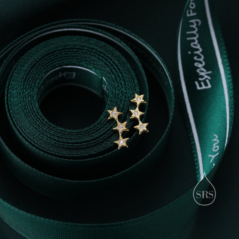 May include: Two gold star-shaped earrings with small clear stones set in each point of the star. The earrings are on a green ribbon with white text that reads "Especially For You".