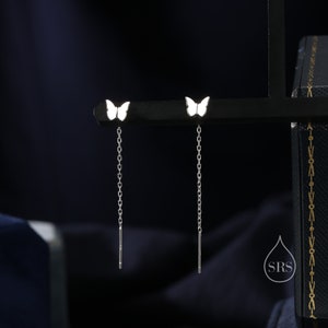 Puede incluir: Un par de pendientes de plata con un dije de mariposa blanca en cada pendiente. Los pendientes tienen una cadena delicada que cuelga del dije de mariposa.