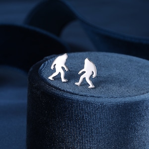May include: A pair of silver earrings featuring a silhouette of a Bigfoot walking. The earrings are displayed on a blue velvet surface.