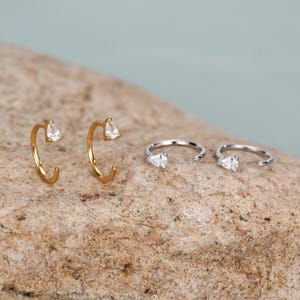 Peut inclure: Une collection de boucles d'oreilles cr&eacute;oles ouvertes avec une pierre transparente en forme de goutte. Deux sont dor&eacute;es et deux sont argent&eacute;es. Les boucles d'oreilles sont pr&eacute;sent&eacute;es sur une surface textur&eacute;e brun clair avec un fond bleu clair.