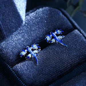 Könnte beinhalten: Ein Paar blaue und weiße Emaille-Ohrringe in Form einer Meeresschnecke. Die Ohrringe haben die Form einer Meeresschnecke mit einem langen Schwanz und mehreren Flossen. Die Ohrringe liegen auf einem blauen Samt-Hintergrund.