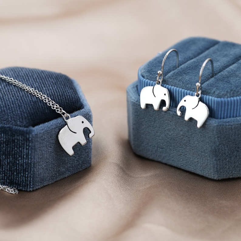 May include: A silver necklace and earrings set with a small elephant charm. The jewelry is displayed on a blue velvet box.
